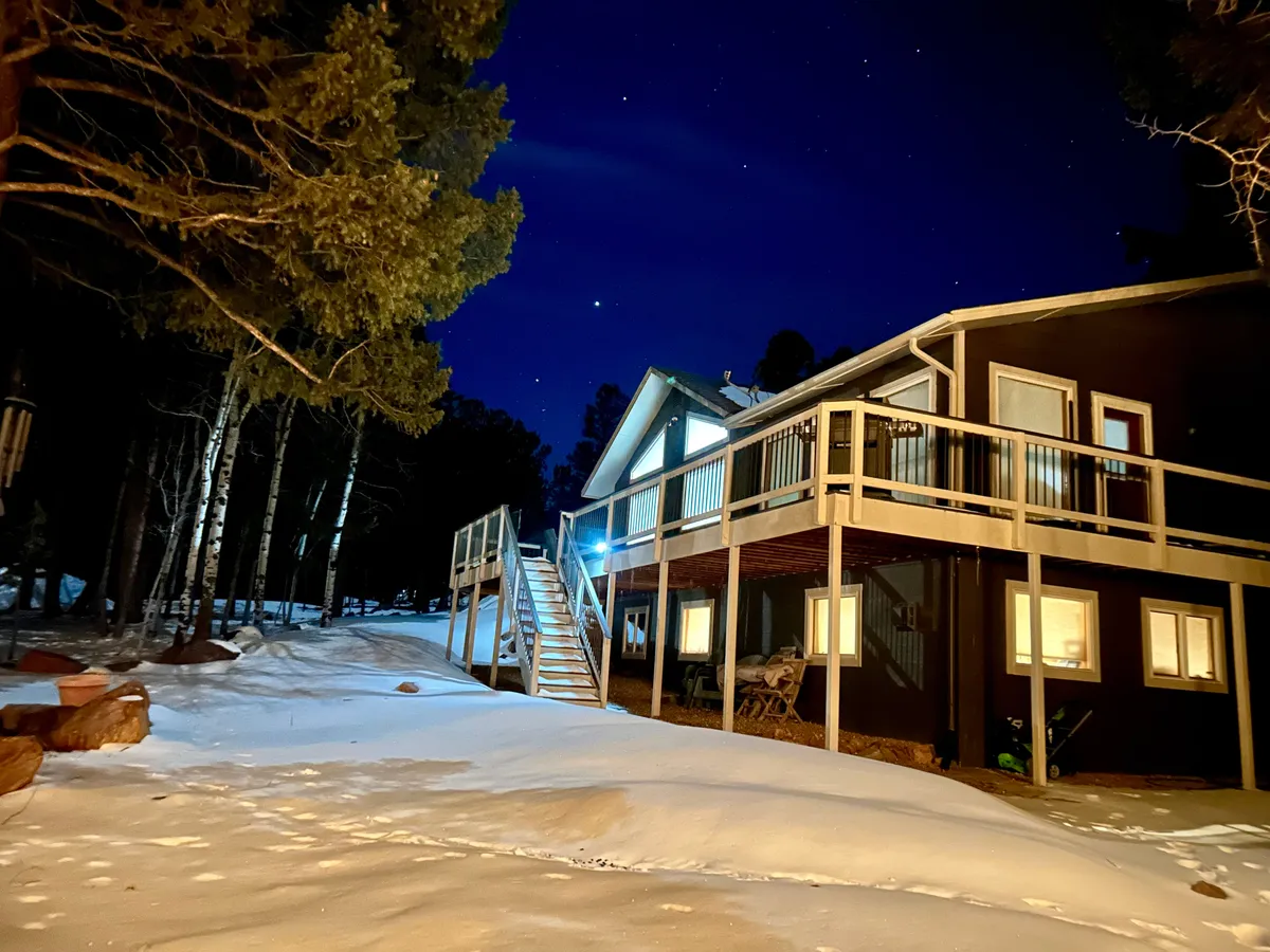 This image features the exterior of the house nestled among snow-covered ground and trees, with a staircase leading up to the home. The setting evokes tranquility, with the property surrounded by dense forest and a cozy, inviting atmosphere. The outdoor space is ideal for peaceful walks or moments of reflection in nature.