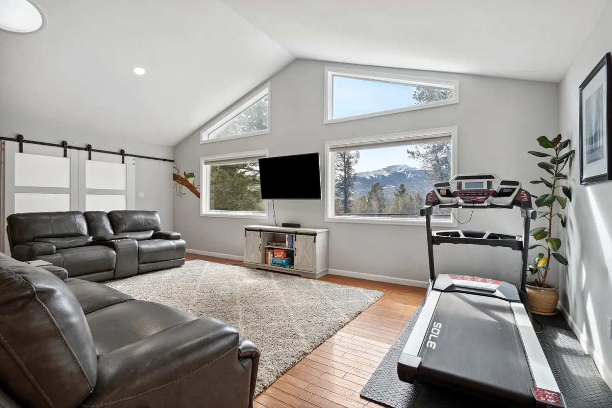 A bright and spacious living area featuring large windows that frame beautiful mountain views. The space includes comfortable seating, a treadmill, and a TV unit, providing a versatile environment for both relaxation and staying active. Sunlight pours into the room, creating a warm and inviting atmosphere.
