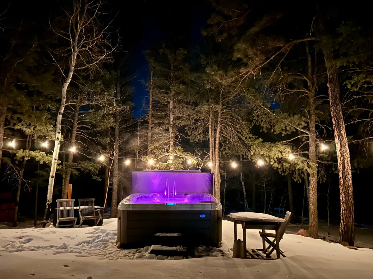 This exterior image captures a hot tub glowing with purple lighting, surrounded by string lights above and snowy ground, with wooden chairs and a table nearby. The ambiance is tranquil and inviting, evoking a sense of cozy relaxation ideal for unwinding after outdoor adventures.