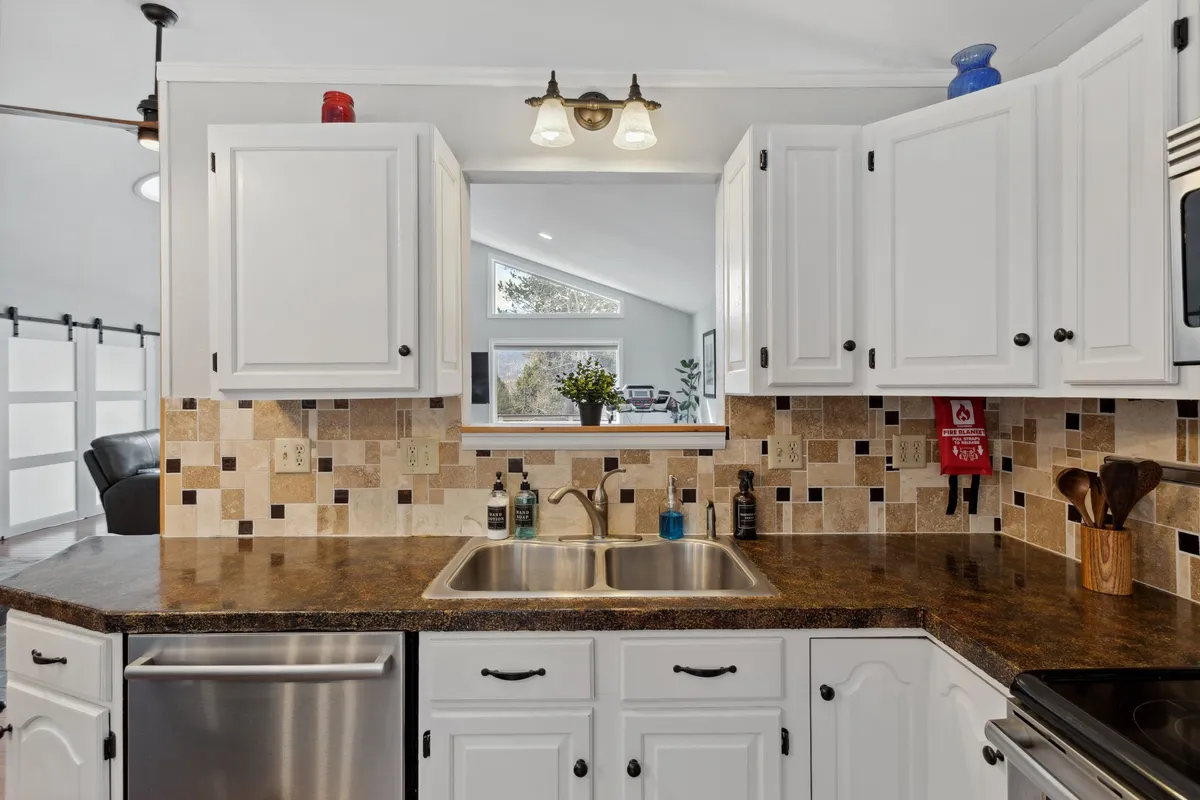 This image showcases a welcoming kitchen with a vibrant, colorful backsplash, a spacious double sink, and a window offering a pleasant view. The scene is infused with natural light, highlighting the modern appliances and open layout, suggesting an ideal place for cooking and entertaining. Family members are gathered, hinting at shared meal preparation and lively culinary moments.