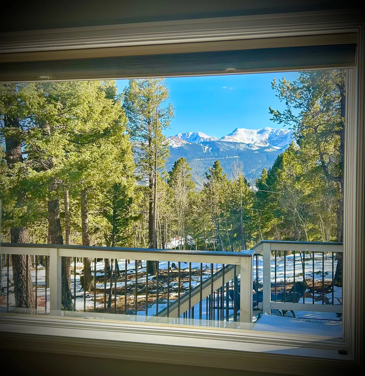 The image showcases a stunning view from the home's deck, overlooking snow-capped mountains and surrounded by tall evergreen trees, with a crisp blue Colorado sky overhead. The scene radiates tranquility and invites guests to immerse themselves in the peaceful natural beauty of Woodland Park.